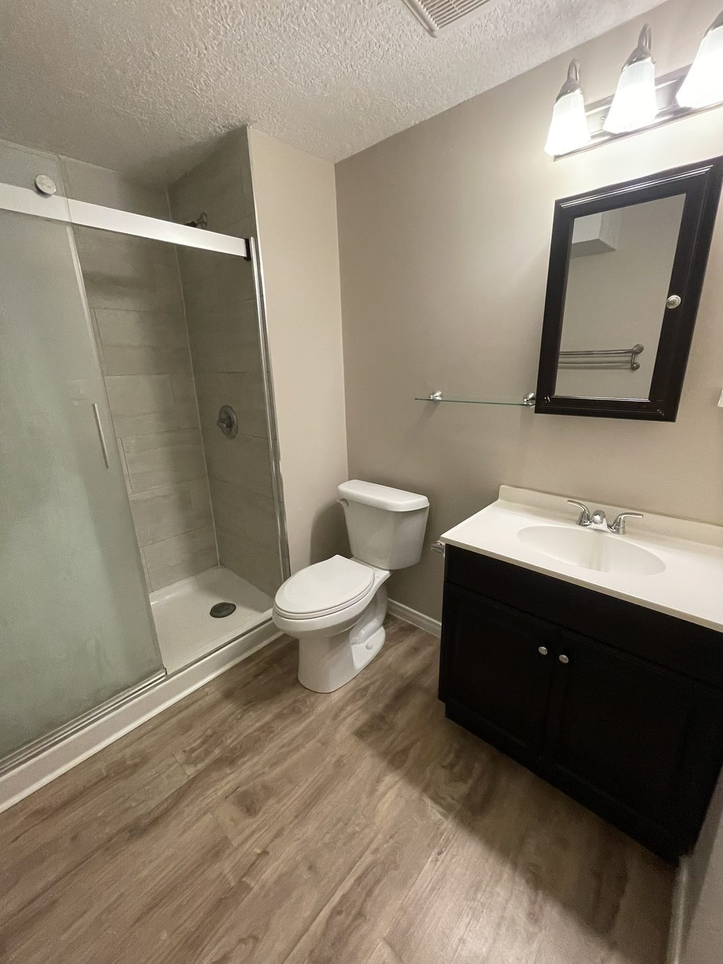 Walk-in glass shower with dark vanity and warm wood-look flooring.